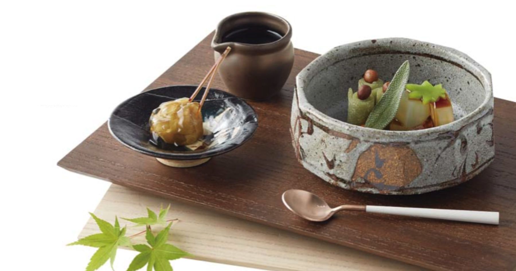 木製のトレイに乗せられた料理。灰色の陶器の器に盛られた野菜と豆、黒い小皿に串刺しの団子、小さな茶色の器に入ったソース。隣に小さなスプーンと箸。