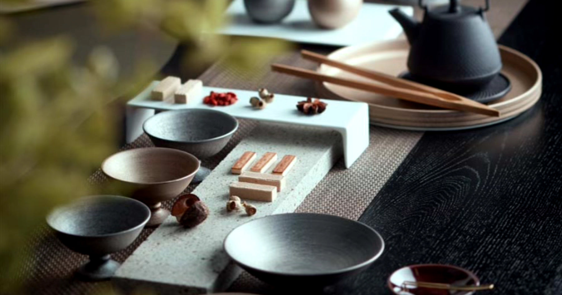 様々な形と色の酒器と小皿がテーブルに並べられている。木製の箸や鉄製の急須も見える。テーブル上にはスパイスや小物が配置され、落ち着いた和風の食卓の雰囲気。背景にぼんやりとした緑の葉。