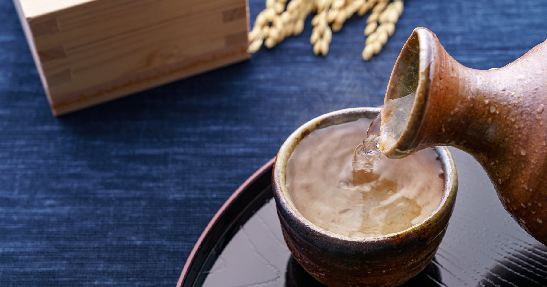 陶器の徳利から盃に日本酒を注ぐ様子。背景には稲穂と木製の箱が置かれている。伝統的な和風の酒器を使用した風景。