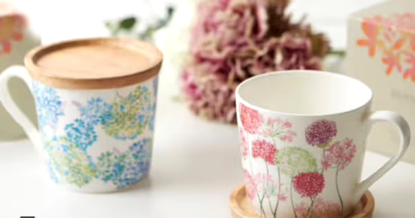 色とりどりの花柄が描かれた2つの白いマグカップがテーブルに置かれている。左のカップは青と緑の花柄で、木製の蓋が付いている。右のカップは赤とピンクの花柄で、木製のコースターに乗せられている。背景にはぼんやりとした花が映っている。