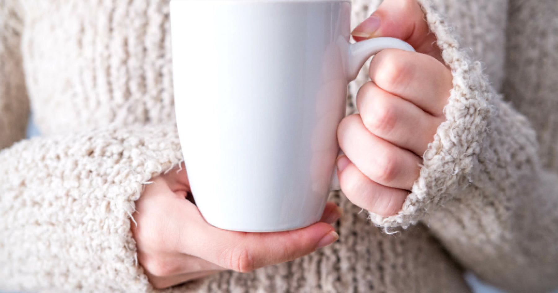 厚手のセーターを着た手が白いマグカップを持っている。寒い季節に温かい飲み物を楽しむ様子。