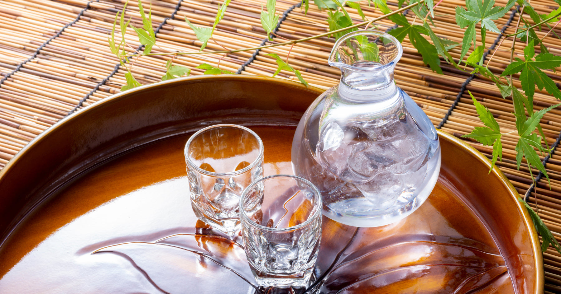 氷を入れた透明なガラス製の徳利と二つのグラスが、竹の敷物の前に置かれた木製のトレイに並べられている。夏の涼しげな雰囲気。背景に青々とした葉。