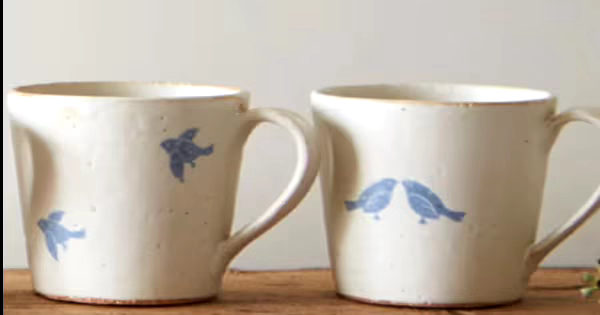 2つの白い陶器のマグカップ。左のカップには青い鳥の絵が描かれ、右のカップには2羽の鳥が向かい合っている絵が描かれている。