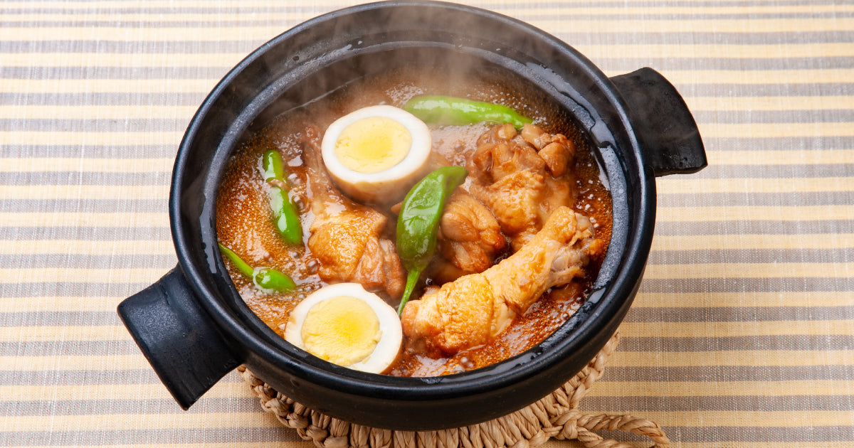 黒い土鍋に盛られた煮込み料理。ゆで卵、鶏肉、そして緑色の唐辛子がスパイシーな赤いスープに浸っている。鍋からは湯気が立ち上り、温かい雰囲気を醸し出している。
