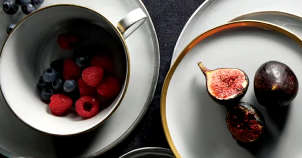 黒い布の上に置かれた白い食器セット。縁に金色の装飾が施されたカップやプレート、ボウルが含まれている。ボウルの中には数粒のぶどうが入っている。