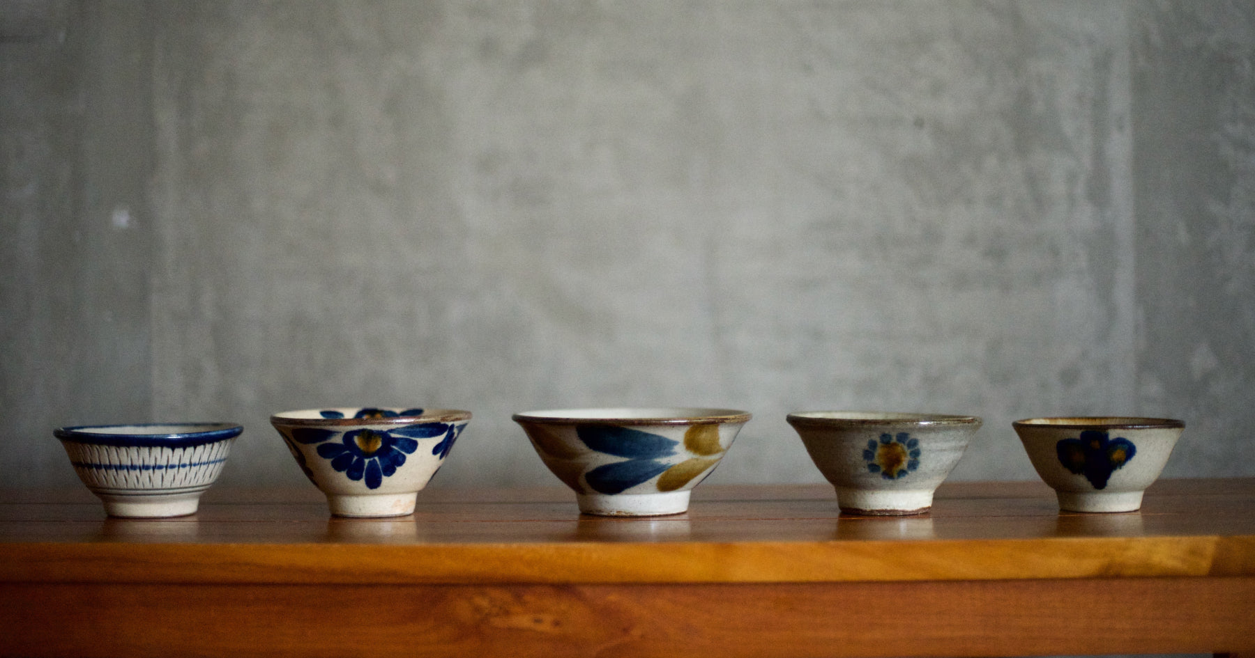 木製のテーブルに並べられた5つの陶器のお茶碗。各碗には異なる青や黄色の花柄が描かれている。背景はグレーの壁。