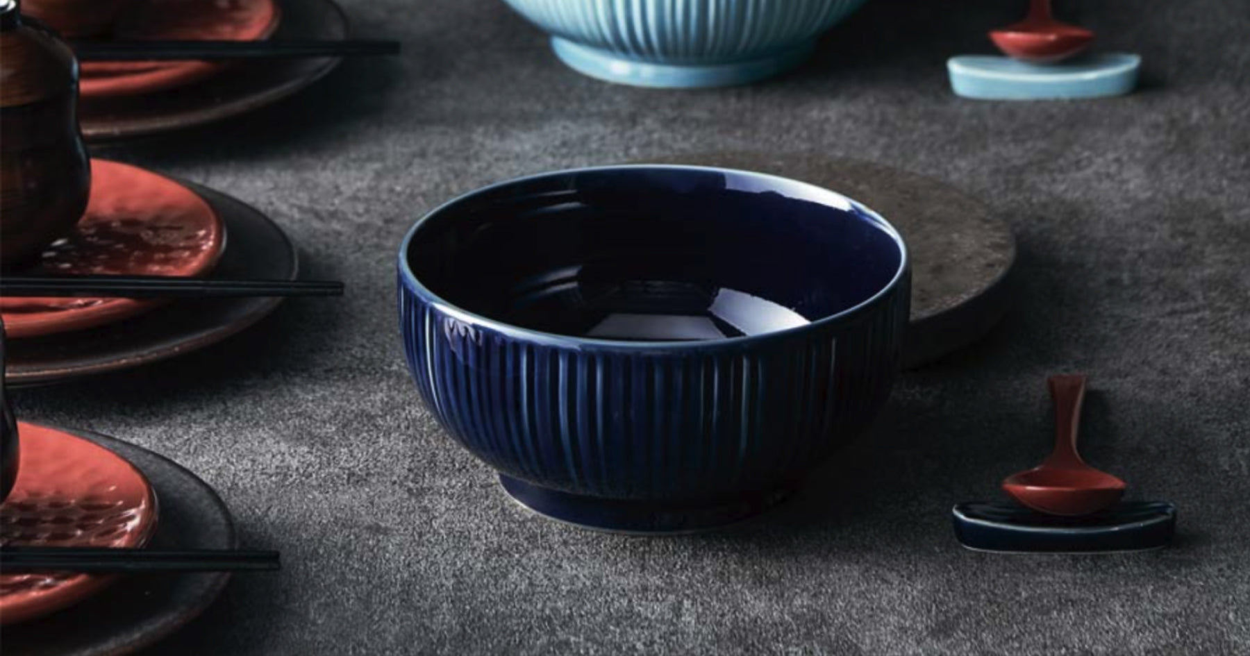 暗い背景に置かれた濃い青色の陶器のお茶碗。隣に赤い小皿と黒い箸が見える。