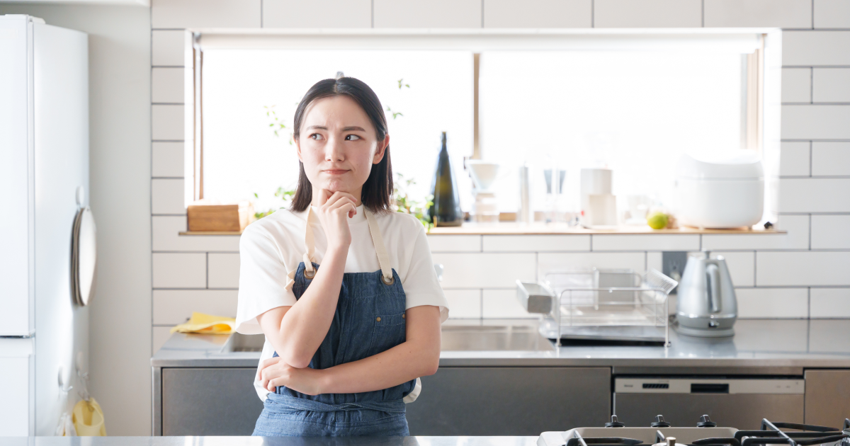 キッチンでエプロンを着け、考え込んだ表情で立っている女性。背景にはシンクや家電製品が整然と配置され、明るい窓から光が差し込んでいる。