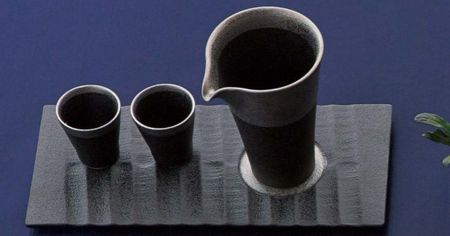 黒い陶器製の徳利と二つの盃が、同じ色合いの長方形のプレートに置かれている。背景は深い青色で、シンプルでモダンなデザイン。
