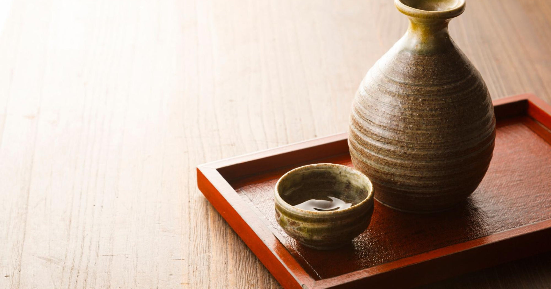 赤い縁の木製トレイに置かれた、陶器製の徳利と盃。木製のテーブルに温かみのある自然な色合い。シンプルで落ち着いた和風の雰囲気。