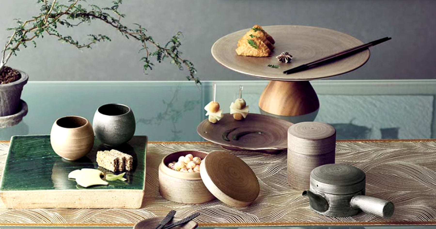 木製の食器や小物が並べられたテーブル。中央には台座付きの大皿があり、料理や箸が置かれている。陶器のカップや蓋付きの容器があり、手前には木製の急須も見える。背景に植物が置かれている。