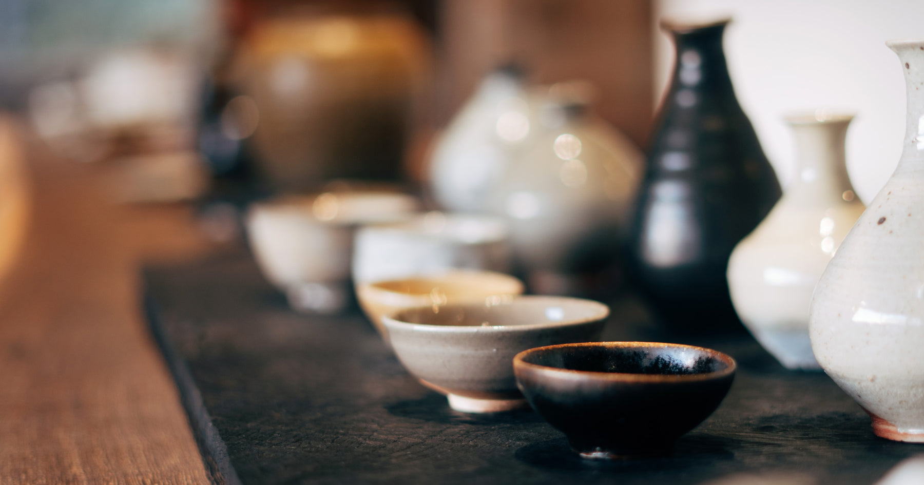 様々な形や色の陶器製の酒器が並べられたテーブル。伝統的な和風の雰囲気。背景にぼんやりとした別の酒器が見える。