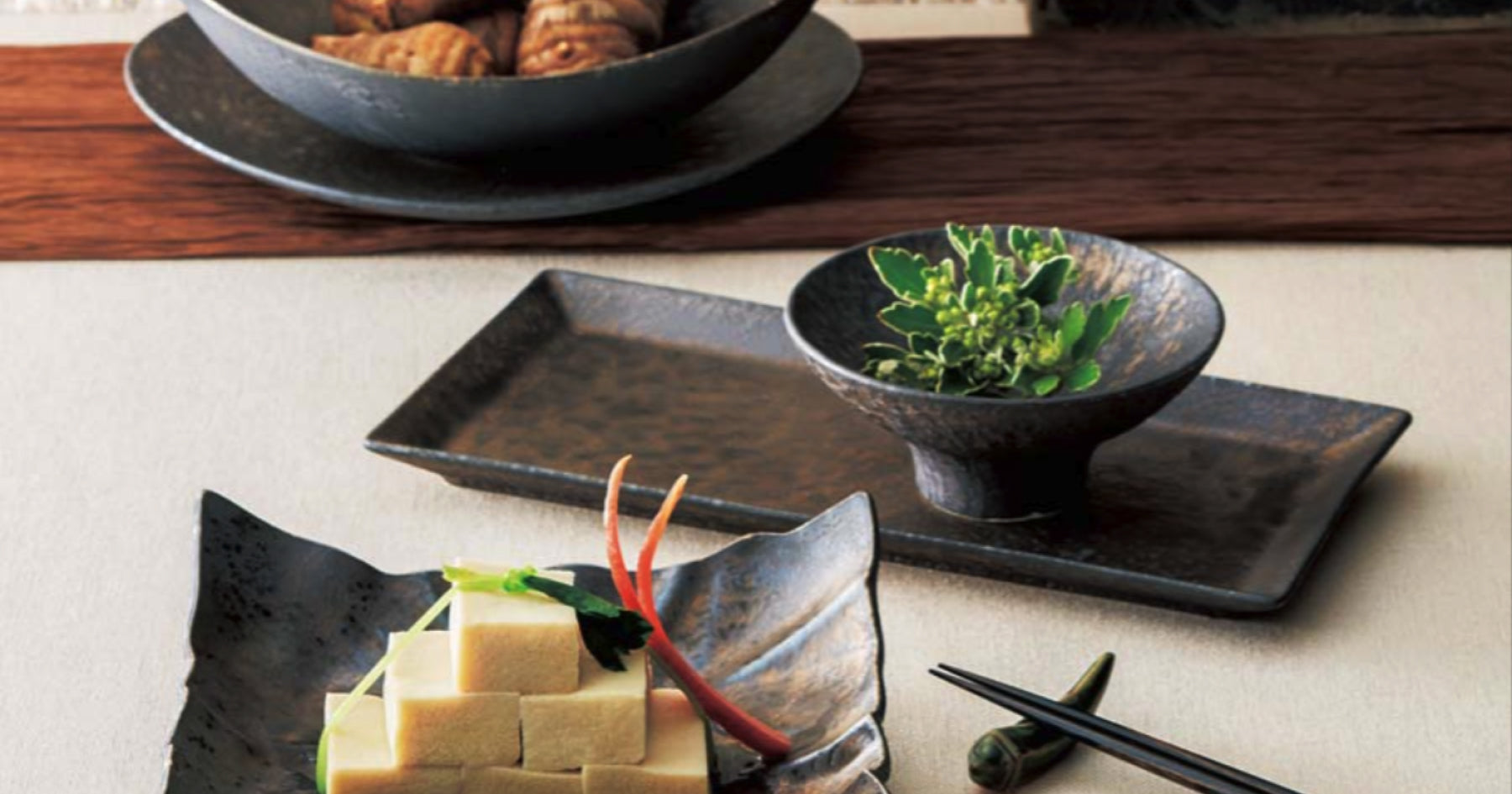 黒い陶器の器に盛られた豆腐料理と野菜、長方形のプレートに乗せられた黒い小鉢に入った緑の葉、背景にある黒い大皿には揚げ物が並んでいる。箸が横に置かれている。
