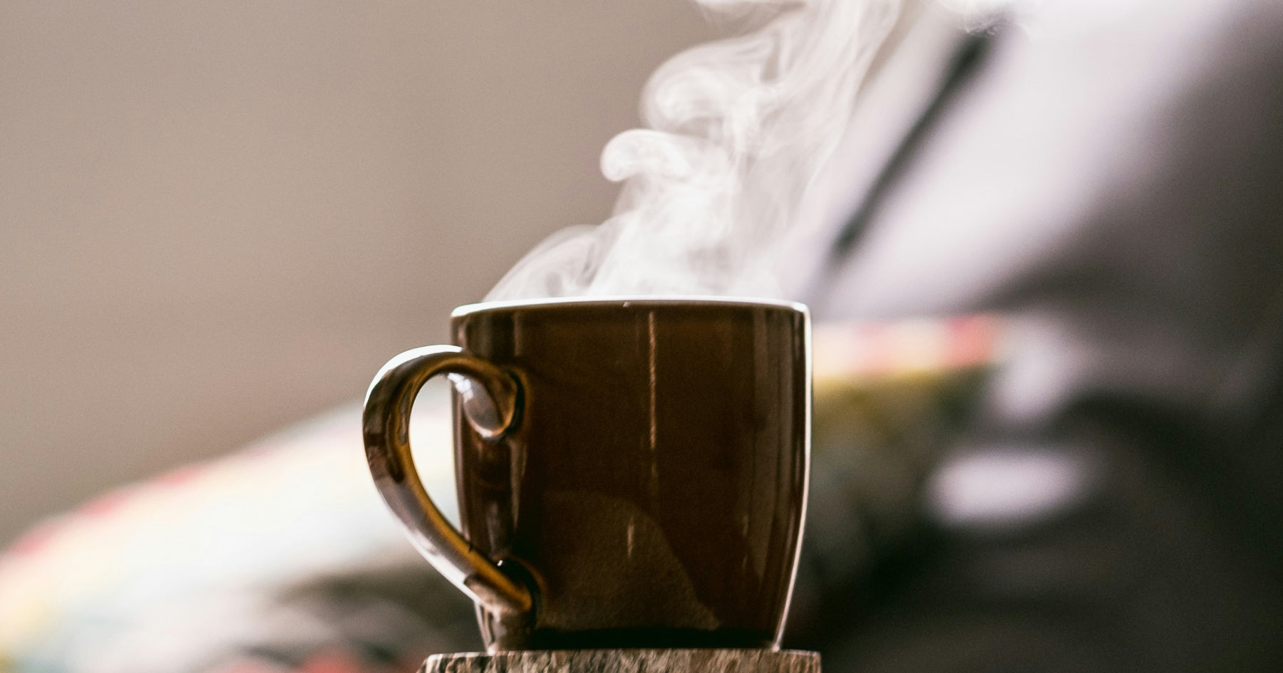 蒸気が立ち上る温かい飲み物が入った茶色のマグカップ。背景はソファやクッションで、リラックスした雰囲気。