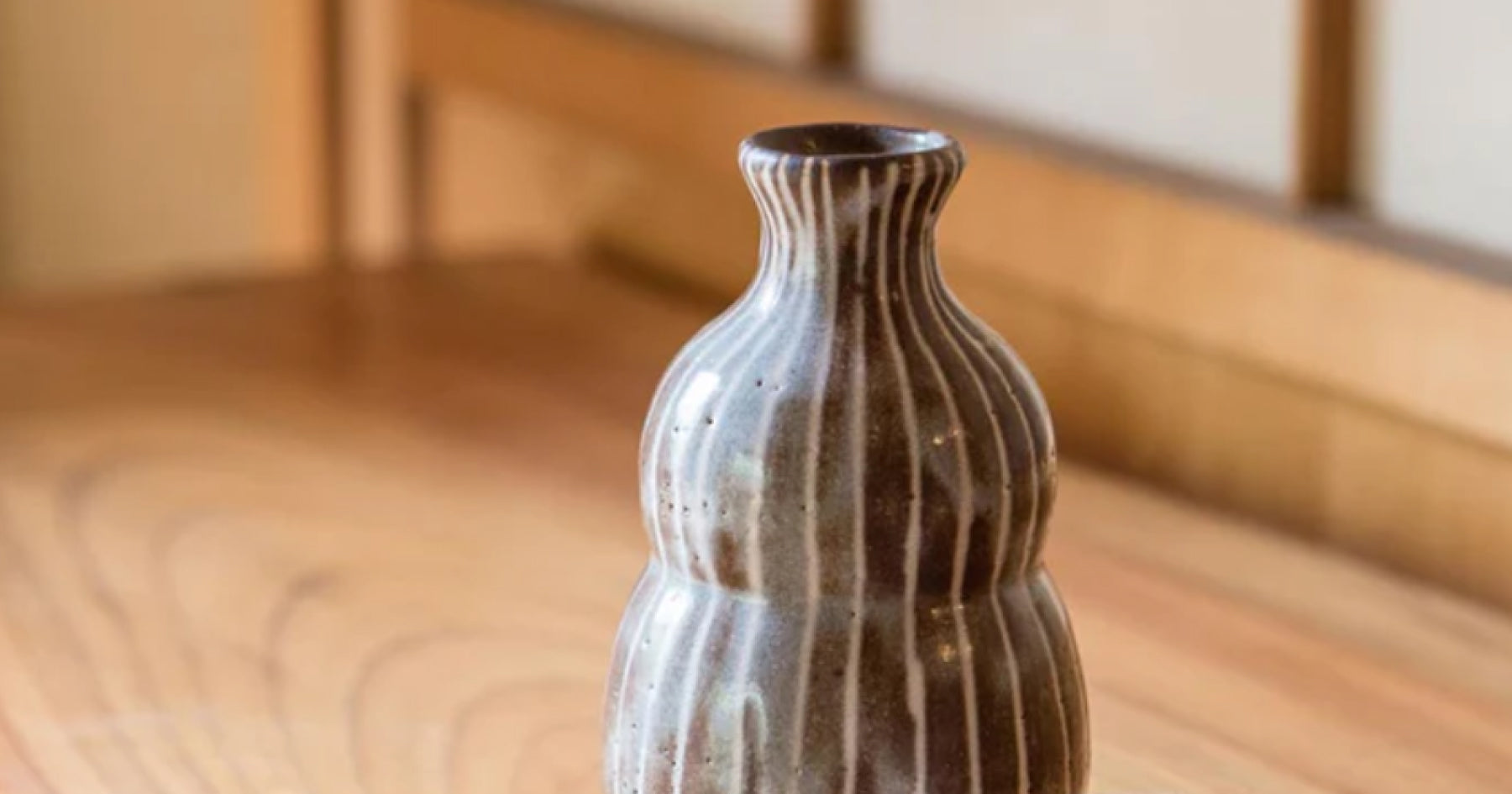 木製のテーブルの上に置かれた、縞模様の陶器製の徳利。背景には和風の室内の一部が見える。シンプルで伝統的な雰囲気。