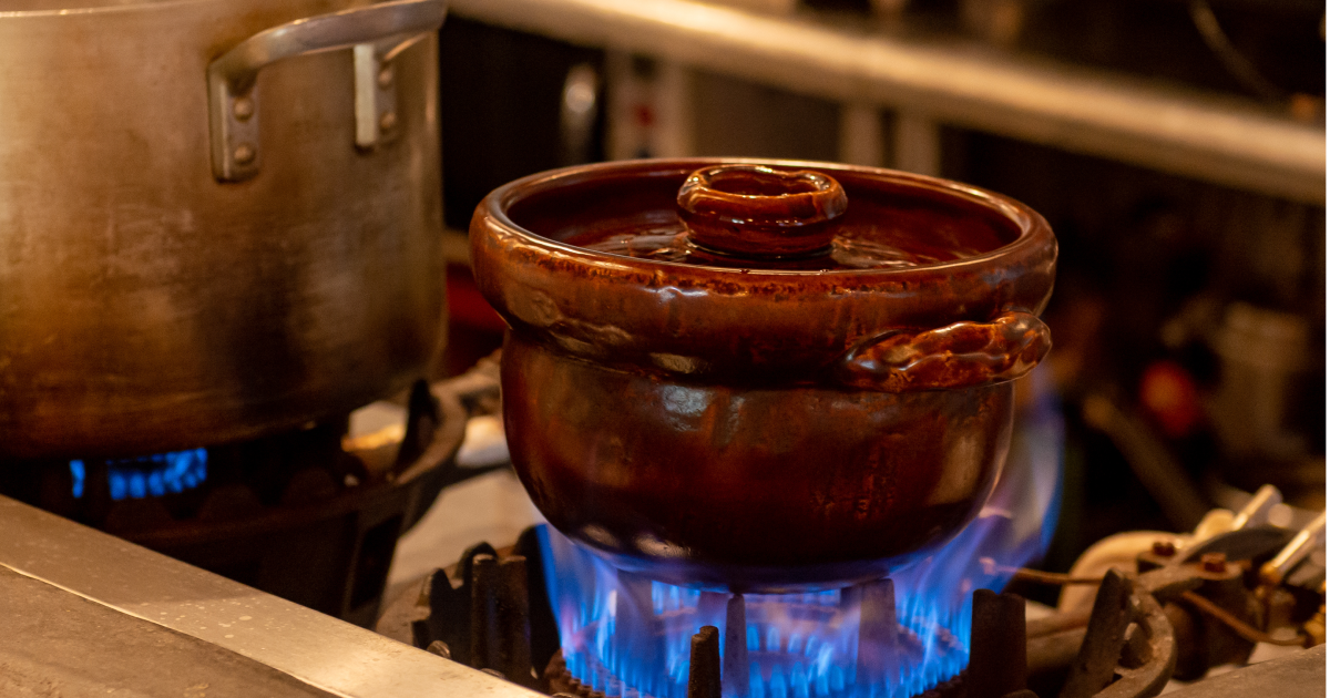 ガスコンロの上で、茶色い陶器の鍋が青い炎で加熱されている。隣には金属製の鍋が同様に加熱されている。厨房の一場面。