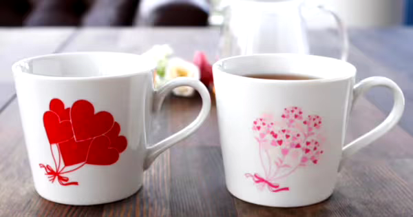 ハートのイラストが描かれた2つの白いマグカップがテーブルに置かれている。左のカップには赤いハートの風船、右のカップにはピンクのハートの花束が描かれている。右のカップにはお茶が注がれている。