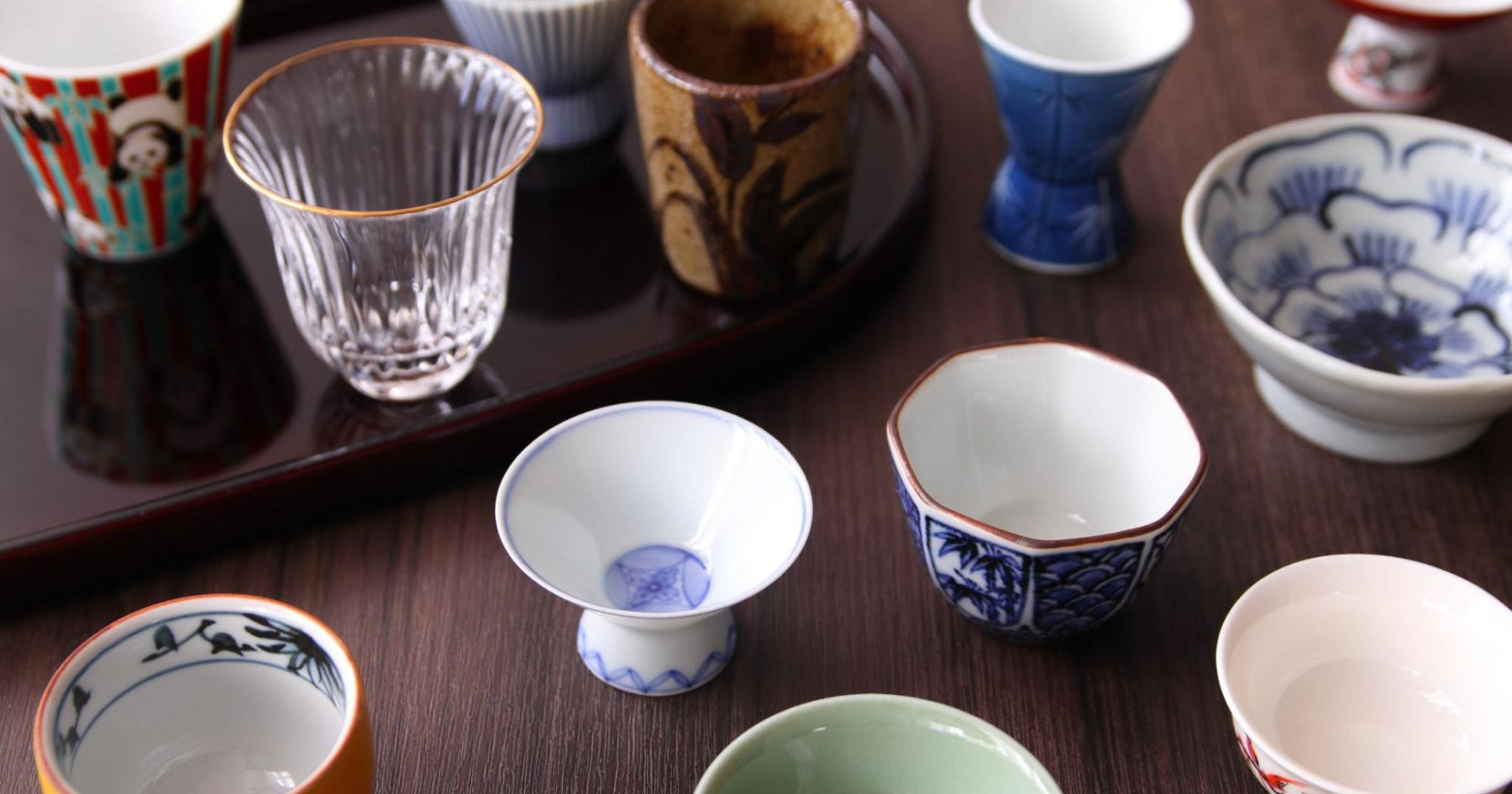 様々な形や色、デザインの酒器がテーブルに並べられている。ガラス製や陶器製の盃が混在し、それぞれに個性的な模様が描かれている。背景には丸い漆器のトレイ。