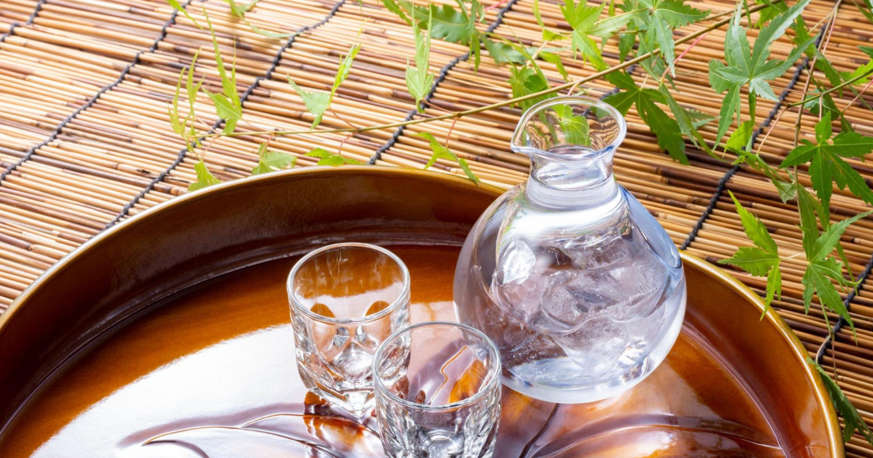 氷を入れた透明なガラス製の徳利と二つのグラスが、竹の敷物の前に置かれた木製のトレイに並べられている。夏の涼しげな雰囲気。背景に青々とした葉。