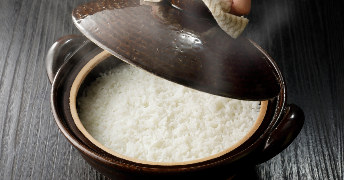 炊きたての白ご飯が入った茶色い土鍋の蓋を開けている場面。湯気が立ち上り、ご飯のふっくらとした質感が見える。