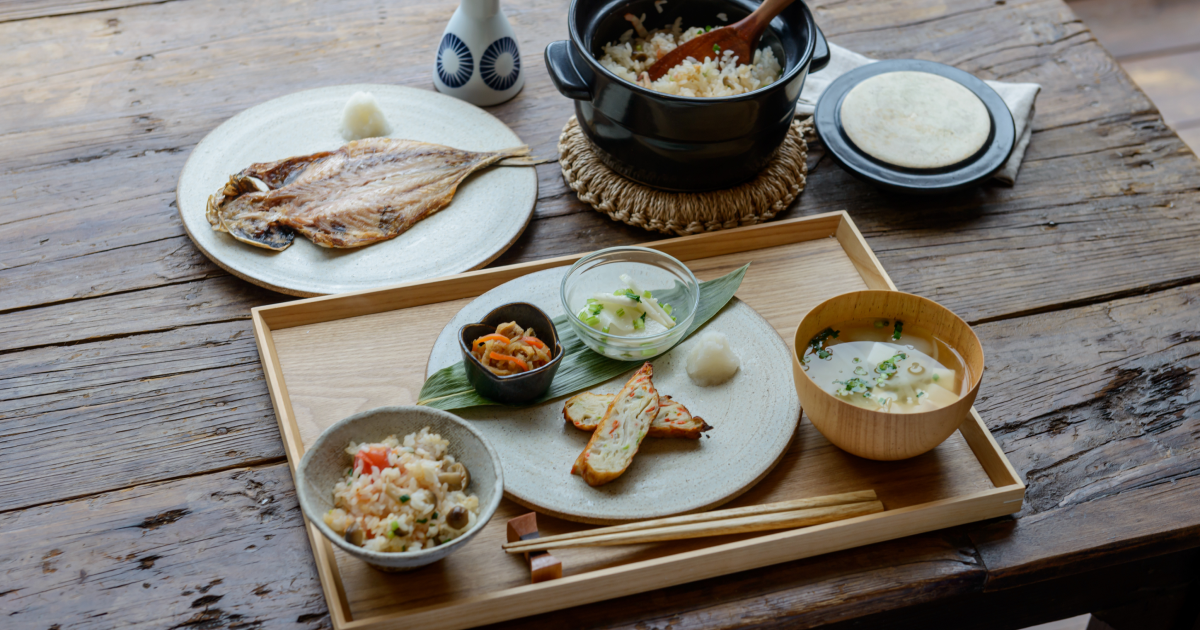 木製のテーブルに並べられた和風の朝食セット。炊き込みご飯、味噌汁、焼き魚、煮物などが陶器の器に盛り付けられている。中央には黒い土鍋に入ったご飯が置かれている。
