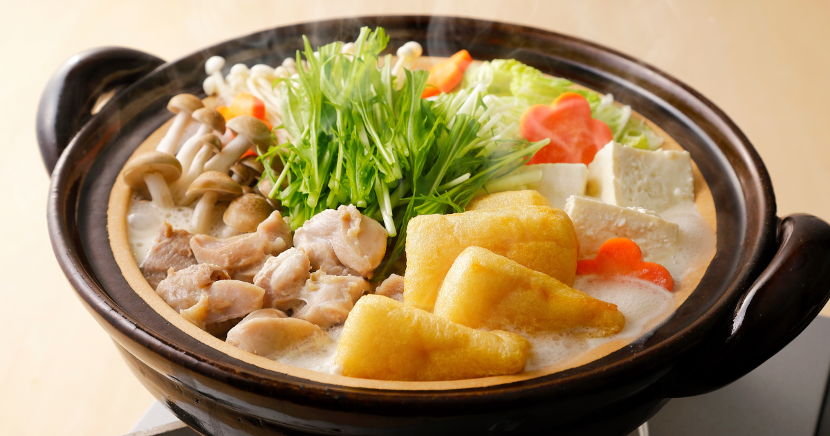 具だくさんの鍋料理が入った土鍋。豆腐、揚げ物、鶏肉、きのこ、白菜、人参、青菜などが盛り付けられており、白いスープが煮立っている。